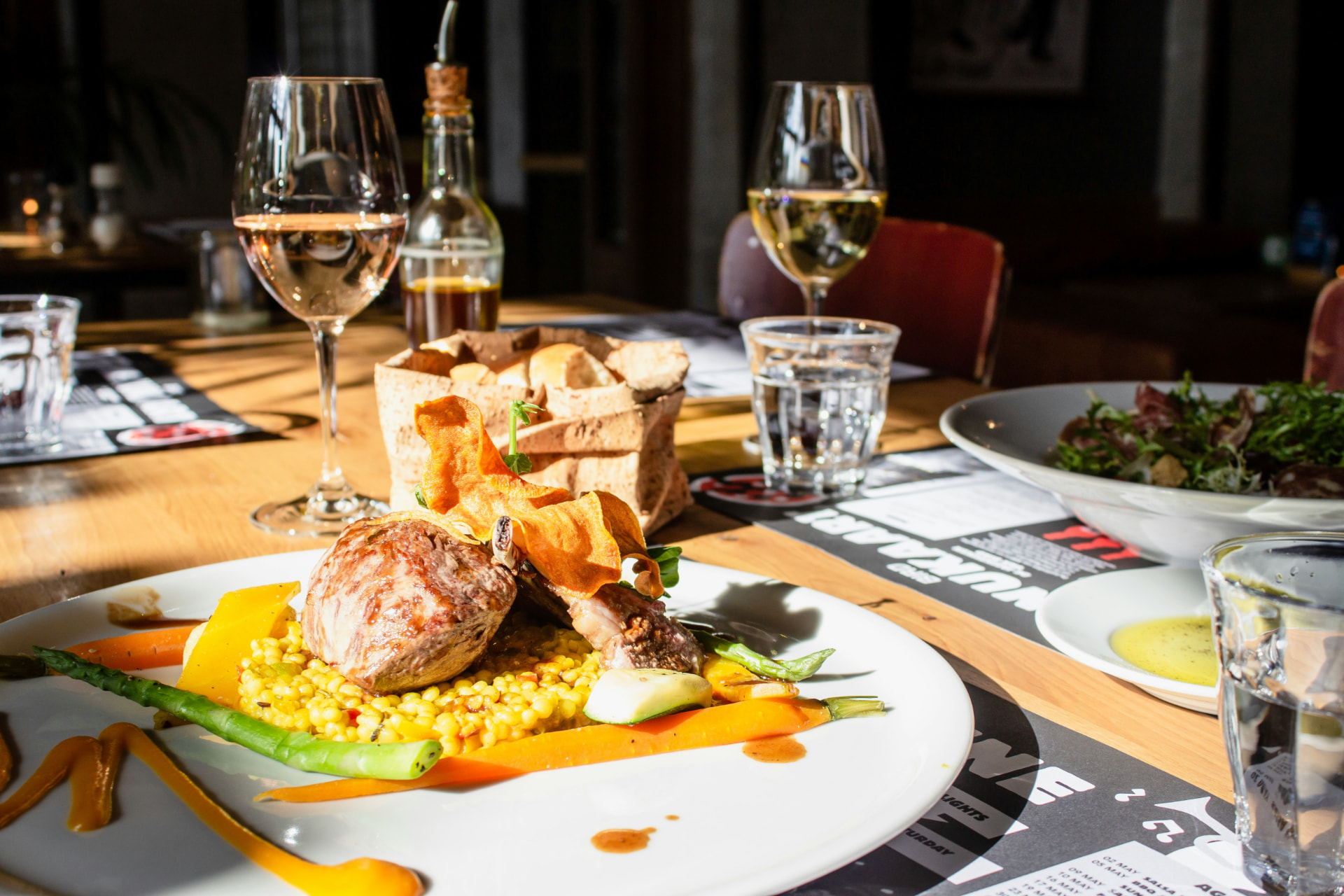 Plated fine dining dish with wine glasses and fresh bread on a sunlit restaurant table