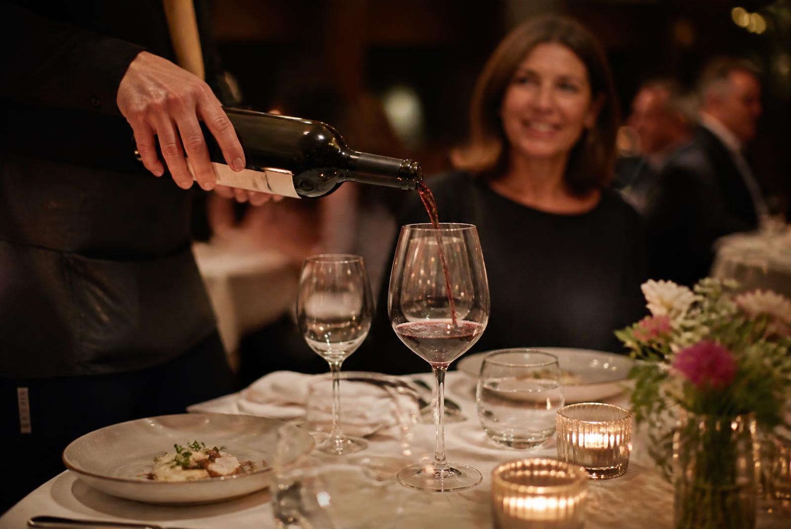 Server pouring red wine for a smiling guest at a candlelit restaurant table