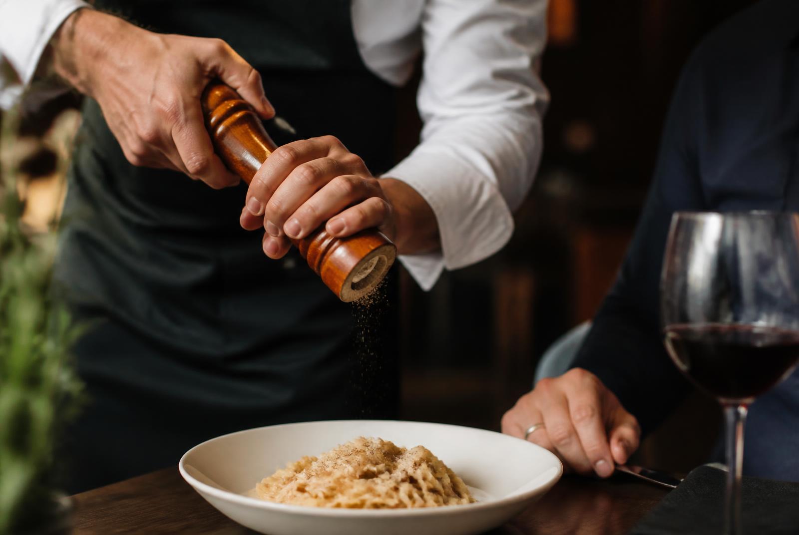 Server grinding fresh pepper over a guest's plated pasta at a restaurant table